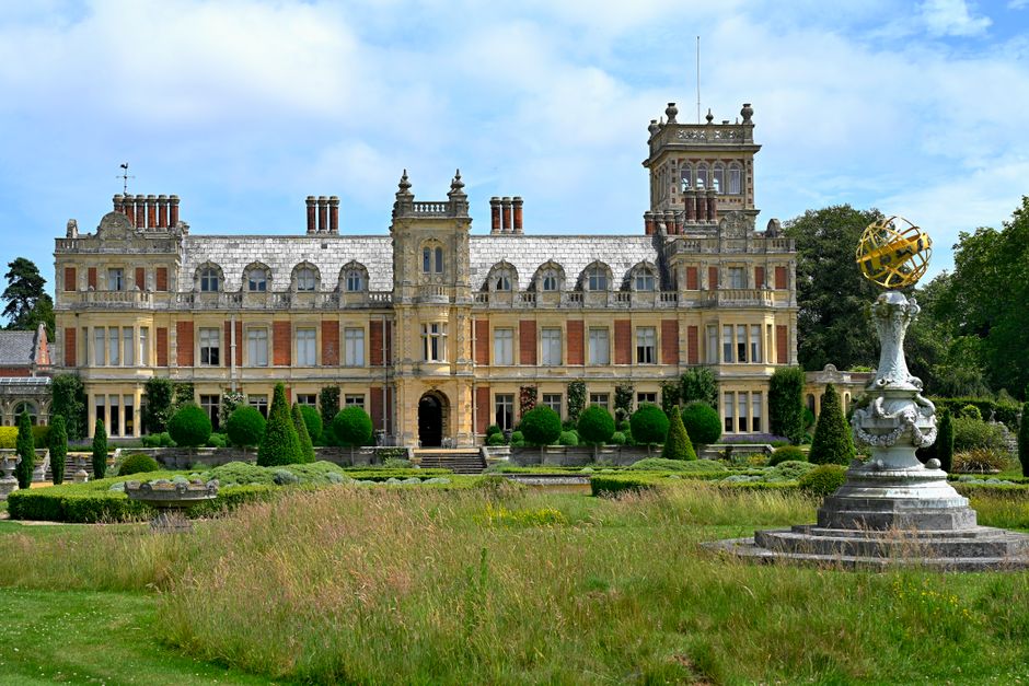 A large, historic mansion with a pale stone facade and multiple ornate towers, set amidst a lush, well-maintained formal garden. The garden features a neatly trimmed lawn and symmetrical flower beds b